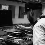 Black and white photo of a DJ using a laptop and mixer indoors, creating music.