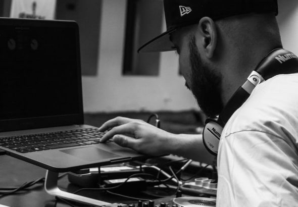 Black and white photo of a DJ using a laptop and mixer indoors, creating music.